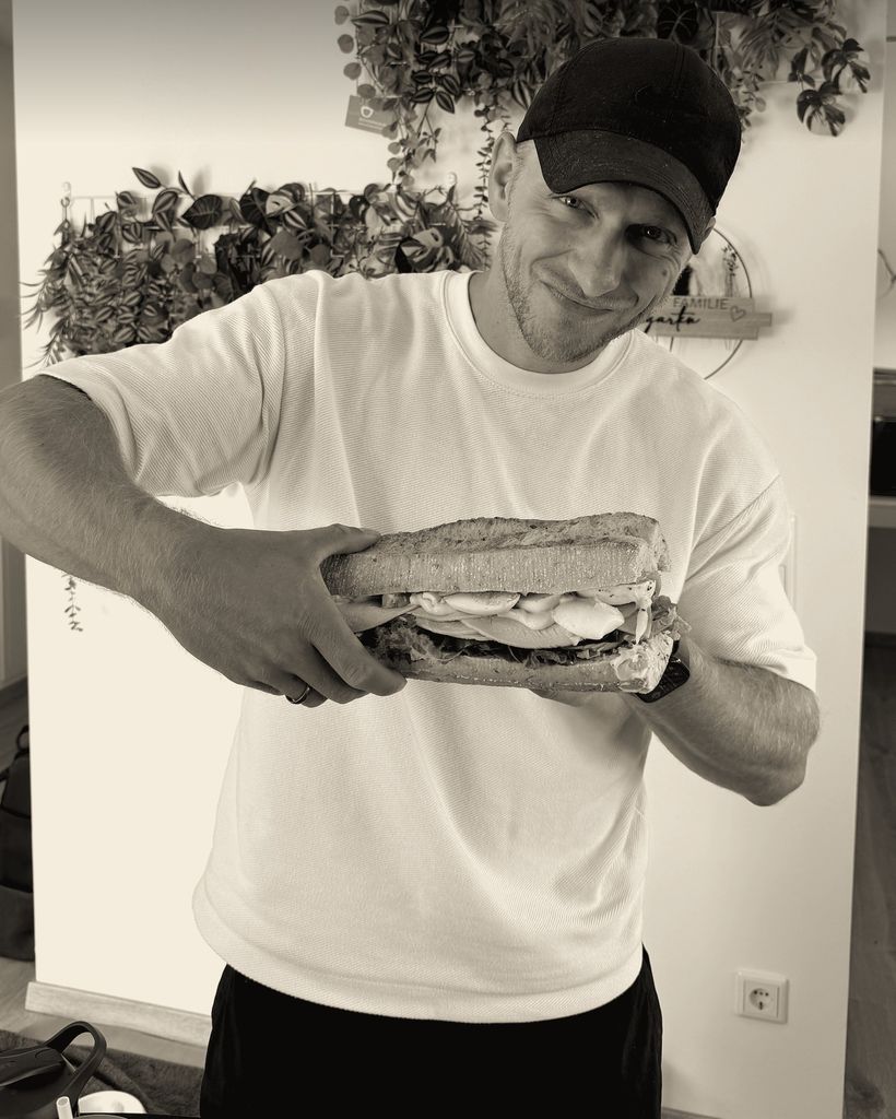 Max grinst und hält ein großes belegtes Baguette in die Kamera.